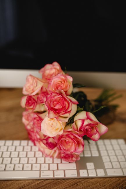 A bouquet of fresh roses with pink and peach petals, arranged in a loose, natural style, resting on a white keyboard placed on a wooden desk. The roses have lush, slightly ruffled petals and green ste
