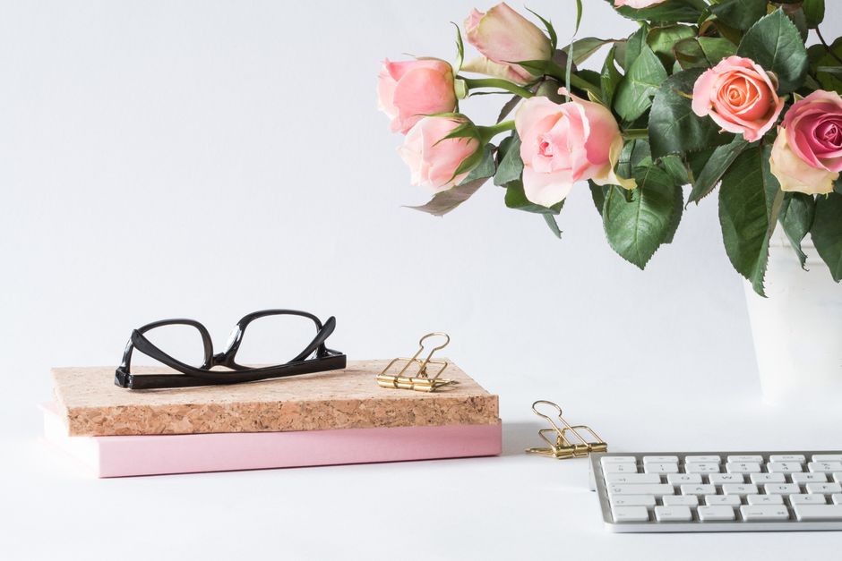 A workspace featuring a bouquet of pink and peach roses with lush green leaves in a white vase positioned on the right side of the image. In the foreground, there is a pair of black eyeglasses resting