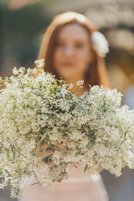 A woman holding a large bouquet of delicate white and light pink flowers, featuring small blossoms and airy foliage, arranged in a loose, natural style. The bouquet is wrapped in light-colored paper o