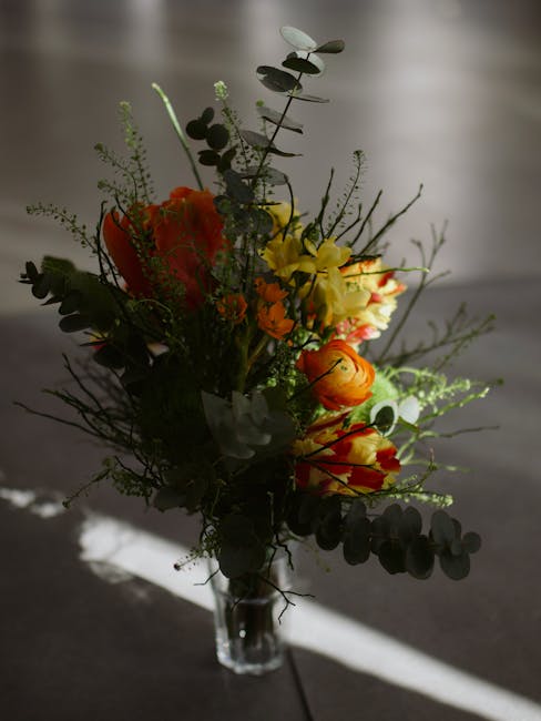 A bouquet of fresh flowers arranged in a clear glass vase. The bouquet features vibrant orange and yellow blooms, complemented by green foliage, including eucalyptus leaves and delicate sprigs of fill