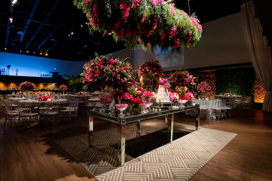 A large floral display featuring vibrant pink roses, lilies, and assorted greenery arranged on a mirrored rectangular table, complemented by smaller matching floral arrangements in glass vases around 