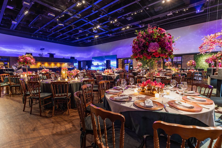 An elegant event space set up for a formal gathering at Broadgate venue, featuring multiple round tables draped with white tablecloths and decorated with large floral centrepieces. The centrepieces ar