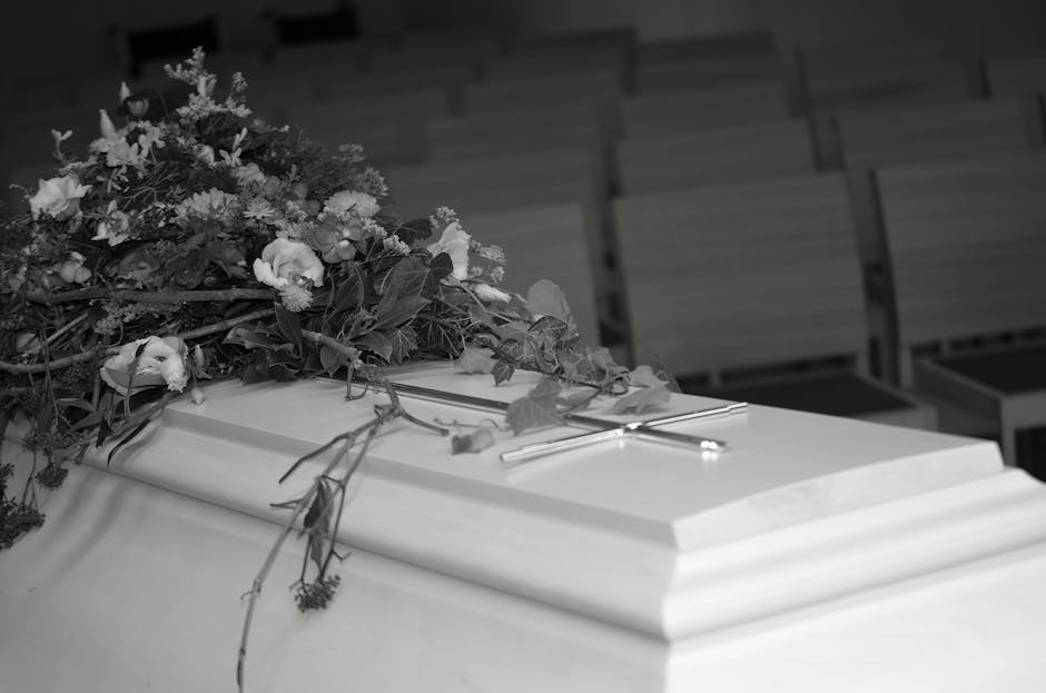 A floral arrangement featuring a lush bouquet of fresh flowers with various blooms and greenery, resting on top of a white coffin with a decorative molding. The bouquet includes petals and leaves with