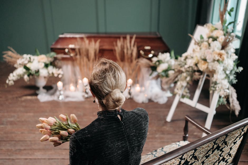 A woman with blonde hair styled in a low bun, wearing a dark velvet dress, is seen from behind holding a bouquet of light pink tulips in her right hand. She is seated on an ornate, wrought-iron and fl