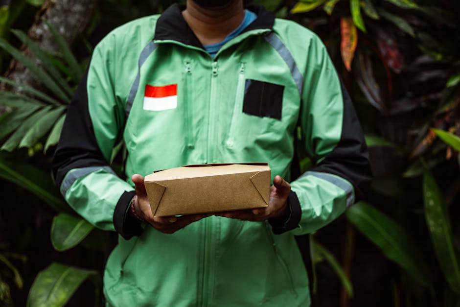 A person wearing a green and black courier jacket with a red and white flag patch on the chest, holding a neatly wrapped brown cardboard parcel with both hands. The background features lush green trop