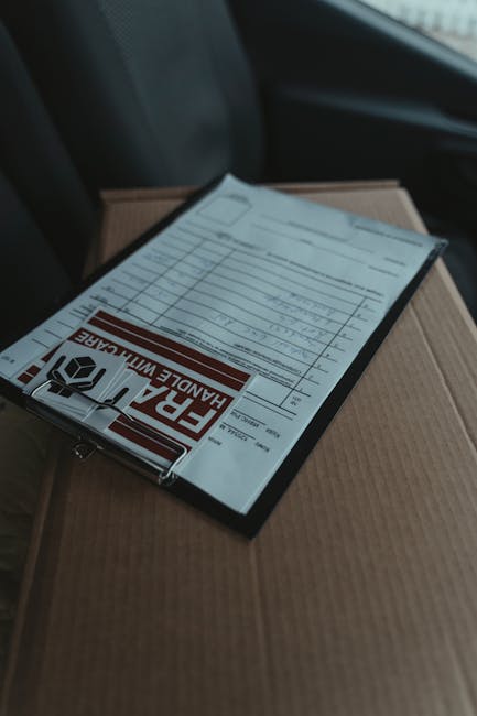 A close-up of a floral gift package resting on top of a cardboard box inside a vehicle. The bouquet features vibrant, fresh flowers including pink roses, white lilies, and purple irises, arranged with