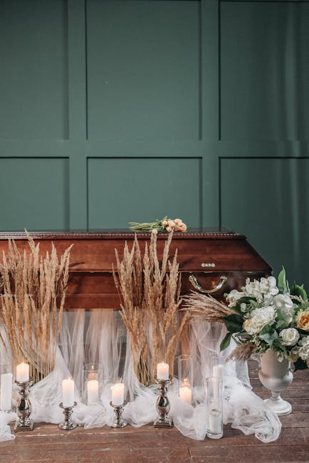 A floral tribute featuring a bouquet of pale pink roses and white blooms resting on a polished wooden casket, surrounded by tall, feathery pampas grass and a variety of white and green flowers arrange