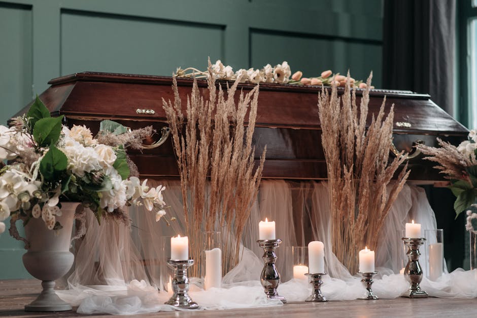 A wooden funeral casket is decorated with a floral arrangement of white roses, carnations, and greenery on top, surrounded by tall beige pampas grass and draped in white fabric. In front of the casket